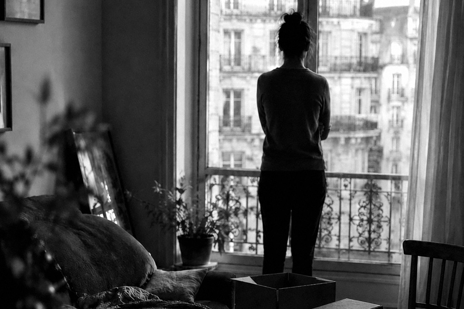 Interior en penumbra de un departamento en el exterior: una mujer de espaldas mira por la ventana hacia la fachada de enfrente, con una caja de mudanza todavía abierta en el piso.
