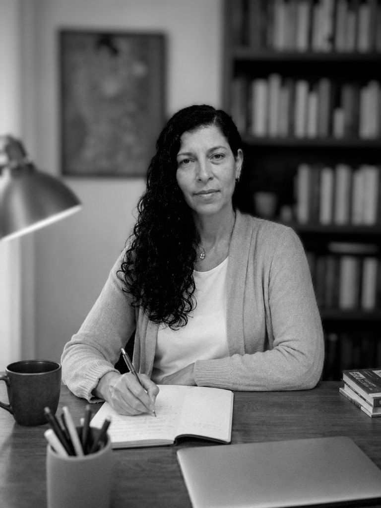 Natalia Fernández, psicóloga, sentada a su escritorio escribiendo en un cuaderno, con una biblioteca detrás.